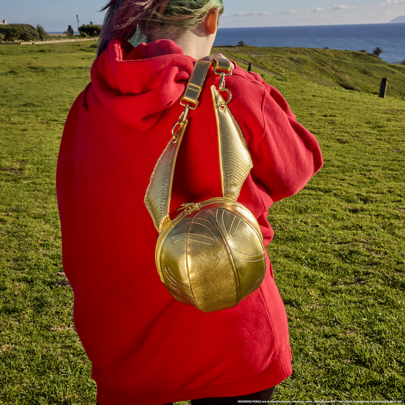Woman wearing a red hoodie on a green field, holding the Snitch crossbody bag over one shoulder 